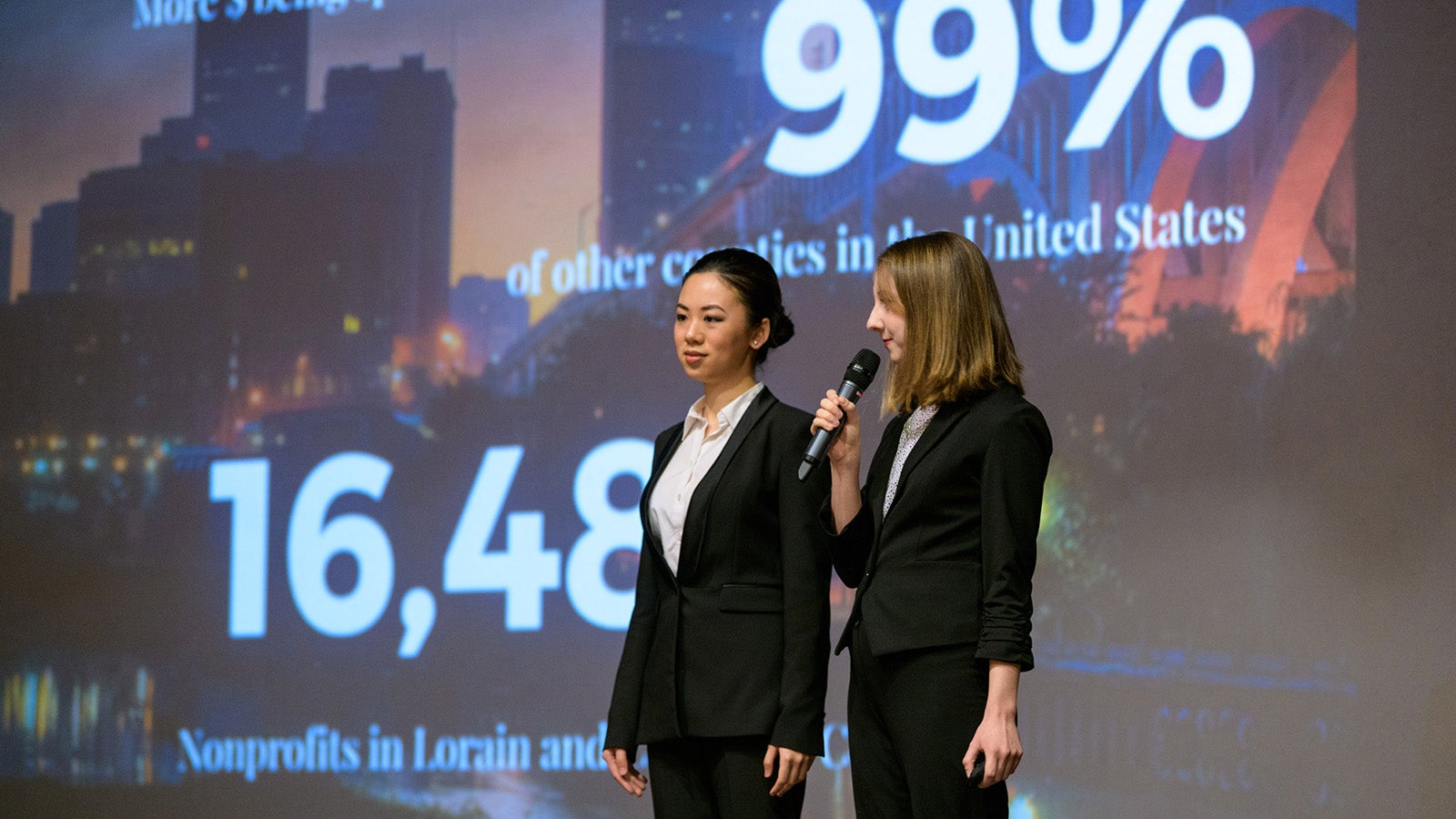 Two students in business attire make a presentation, with numbers projected behind them.