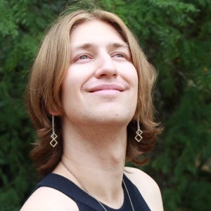 A picture of Evan Hamilton with their hair down and geometric earings on, staring upward against a backdrop of trees.