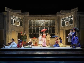 A stage performance featuring actors dressed in period clothing holding colorful umbrellas, posed in a tableau reminiscent of a park scene, with painted animal cutouts and a large, weathered architectural backdrop with grid-like windows.