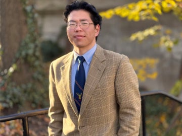 a person wearing glasses, a tan suit jacket and light blue shirt stands outside