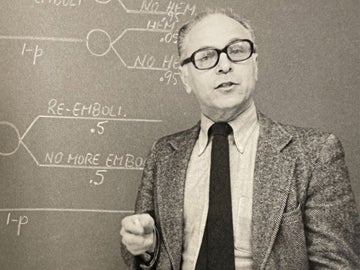 a person wearing glasses stands next to a chalkboard with math equations