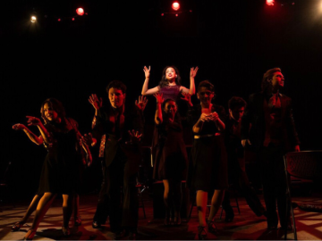a group of performers are gathered around a young woman with a spotlight on her face and her arms up