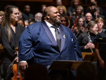 Limmie Pulliam at a podium, cheerfully looking at the audience with an orchestra behind him.