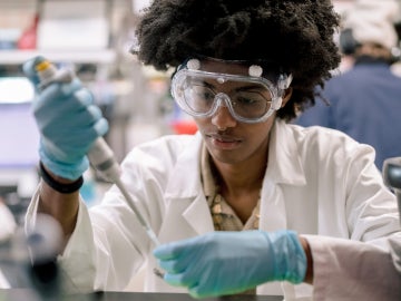 a student wearing a lab coat and blue gloves works in a lab with a pipette