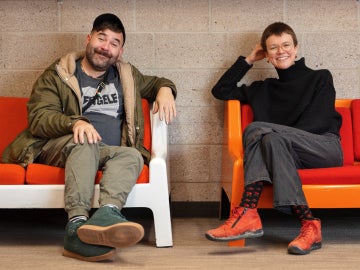 two smiling people sitting on couches with orange pillows