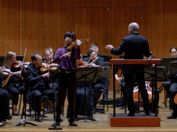 Young man in purple shirt and black pants plays violin on stage in front of full orchestra
