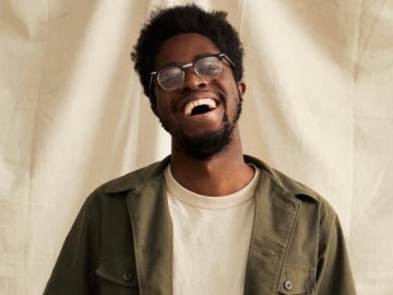 a person with short hair and glasses wearing a white shirt under an olive jacket who is laughing and smiling