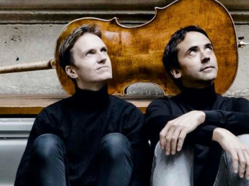 two men wearing black seated together in front of a cello.