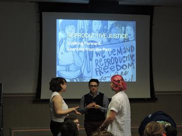 Three people stand in front of a screen with a projected image. The text reads "Reproductive Justice, Looking Forward, Learning from the Past."