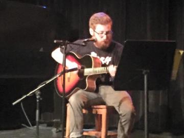 A guitarist is seated by microphones and a music stand