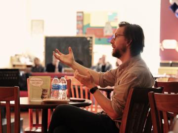 Chris Eldridge, seated at a cafe table, gesturing towards a music ensemble off-frame.
