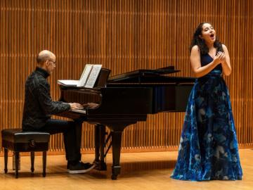 young woman standing in blue gown singing with man in black sweater and pants playing piano