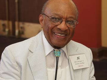 black man with glasses, dressed in beige-striped suit and smiling.