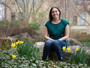 young woman in green shirt and jeans sits on a rock with daffodils.