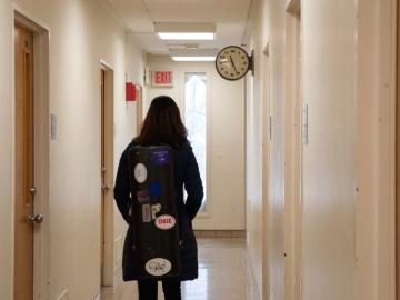 A student walks down a hallway, carrying an instrument case covered in stickers.