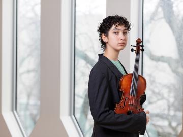 violinist wearing gray suite and with short curly hair stands in front of windows