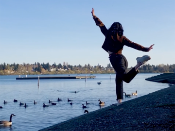 student in brown jacket and dark pants dances in front of a pond with ducks.