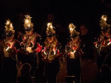 A marching band performs indoors, in the dark, with lights shining from their hats and uniforms.