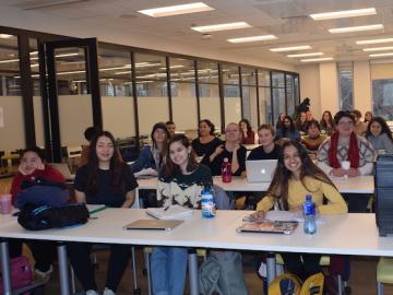 Students sitting at long tables with computers and laptops in a classroom
