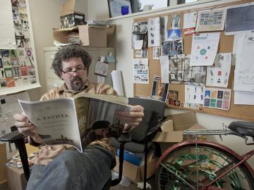 Man with glasses sitting in chair office, reading a magazine, next to a bicycle