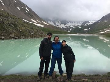 Three people standing in front of mountains and lake. Photo.