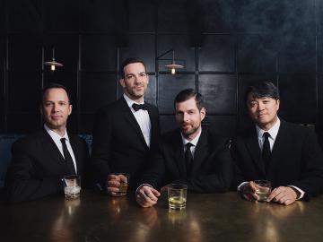 Four men in black suits seated at a bar