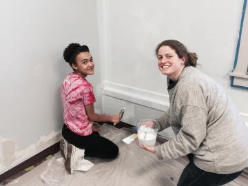 A girl and a young woman paint a baseboard together.