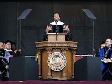Michelle Obama speaks at a podium bearing the Oberlin College seal. Dignitaries in academic robes are seated on both sides.