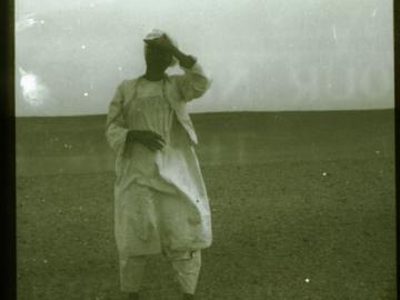 A man in white clothing stands alone in the foreground, an empty landscape behind him. Historical black & white photo.