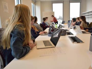 Photo of a students sitting around large desk watching professor write notes in front of class