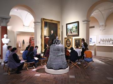 Students look at paintings in the museum while seated in chairs in the gallery.