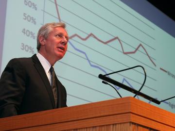 Jim Margolis at a speaking podium. A line chart is projected on the screen behind him.