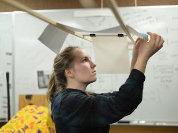 A student inspects a sheet of paper that is draped over 2 wooden rods above her..