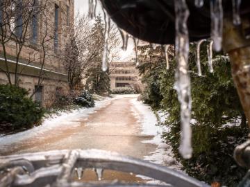 An icicle hangs from a bike seat in the foreground. Behind, a snowy path leads to a building.