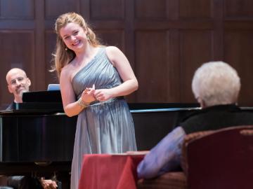 A singer, standing by a grand piano, awaits feedback from Marilyn Horne, who is seated at a small table.