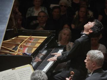 A pianist performs before an audience. He is leaning back with eyes closed and fingers on the keys.
