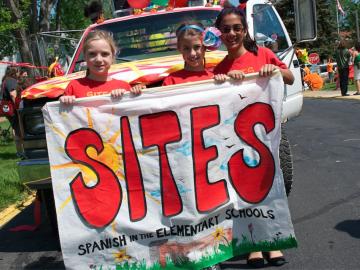 3 school-age girls hold a sign that reads SITES Spanish in the Elementary Schools
