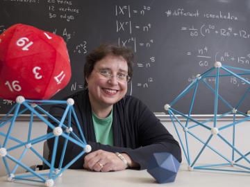 Professor Colley with geometric models on her desk. The blackboard behind her is filled with equations.