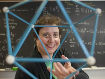 Susan Colley holding a geometric model. In the background, a chalkboard with equations.
