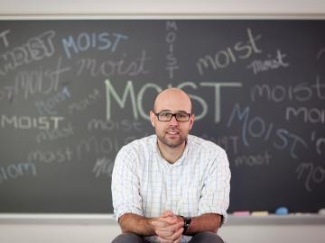 Man seated in front of chalkboard showing the word 'moist'