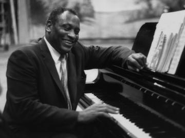Paul Robeson seated at a piano, sharing a laugh with someone. Black and white photo.
