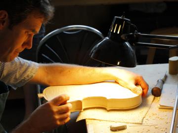 Under a bright lamp, a man works on unfinished wood in the shape of a violin body