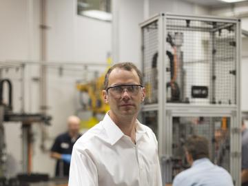 A man in a large workshop wears safety goggles and a white dress shirt. Behind him are two workers and a metal cage containing an apparatus. 