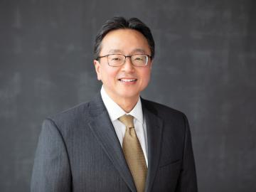 David Kamitsuka smiling with glasses gray suit coat and tan tie.