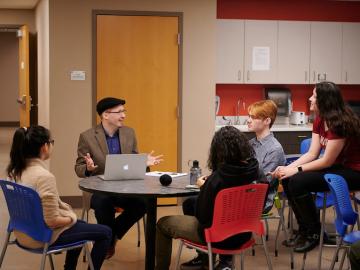 Four students and professor talk while seated at a round table.