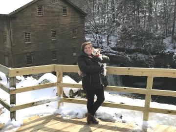 A woman holds a small dog while standing on a wooden ramp near a barn.