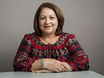 A portrait of a middle-aged woman wearing with arms resting on a table.