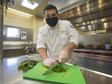 A man wearing a chef jacket chops parsley on a cutting board.
