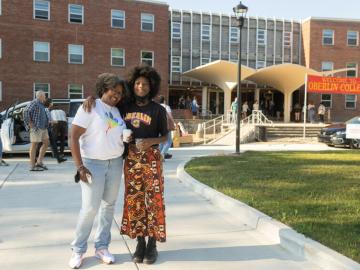 A mother, left, stands in a hug with her student, right, in front of a residence hall. The student is wearing an Oberlin sweatshirt. An Oberlin welcome sign hangs in the background.