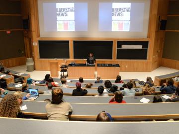 Lecture hall during a presentation. The words 'Breaking News' are visible on the screen behind the lecturer.